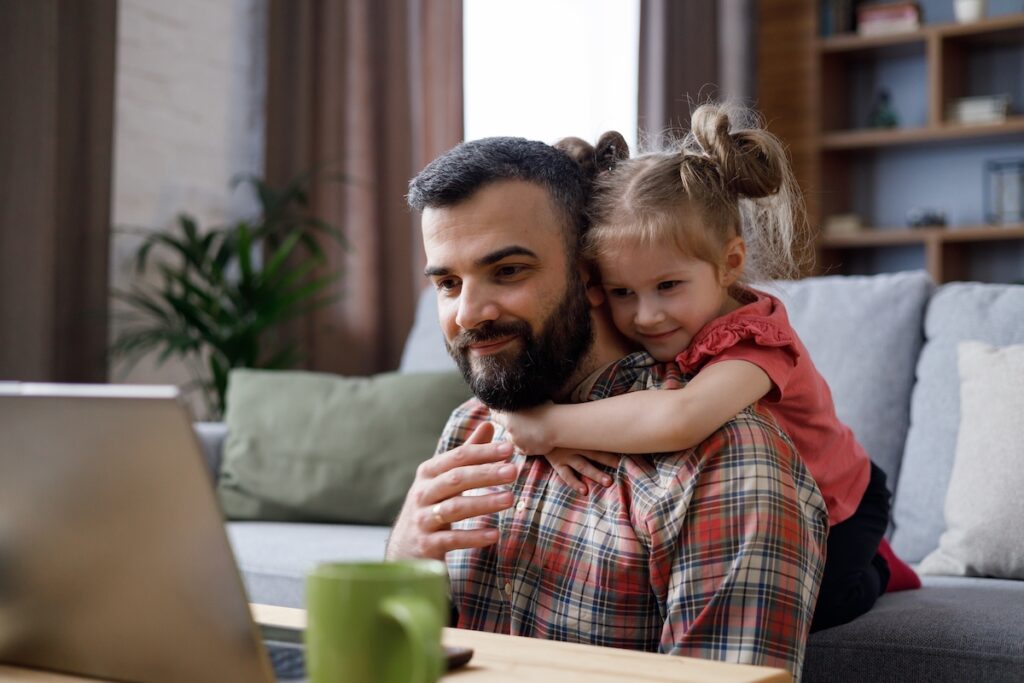 Happy family portrait. Young handsome man works from home at laptop, focus attention on laptop screen while his little daughter hugging her beloved dad around neck